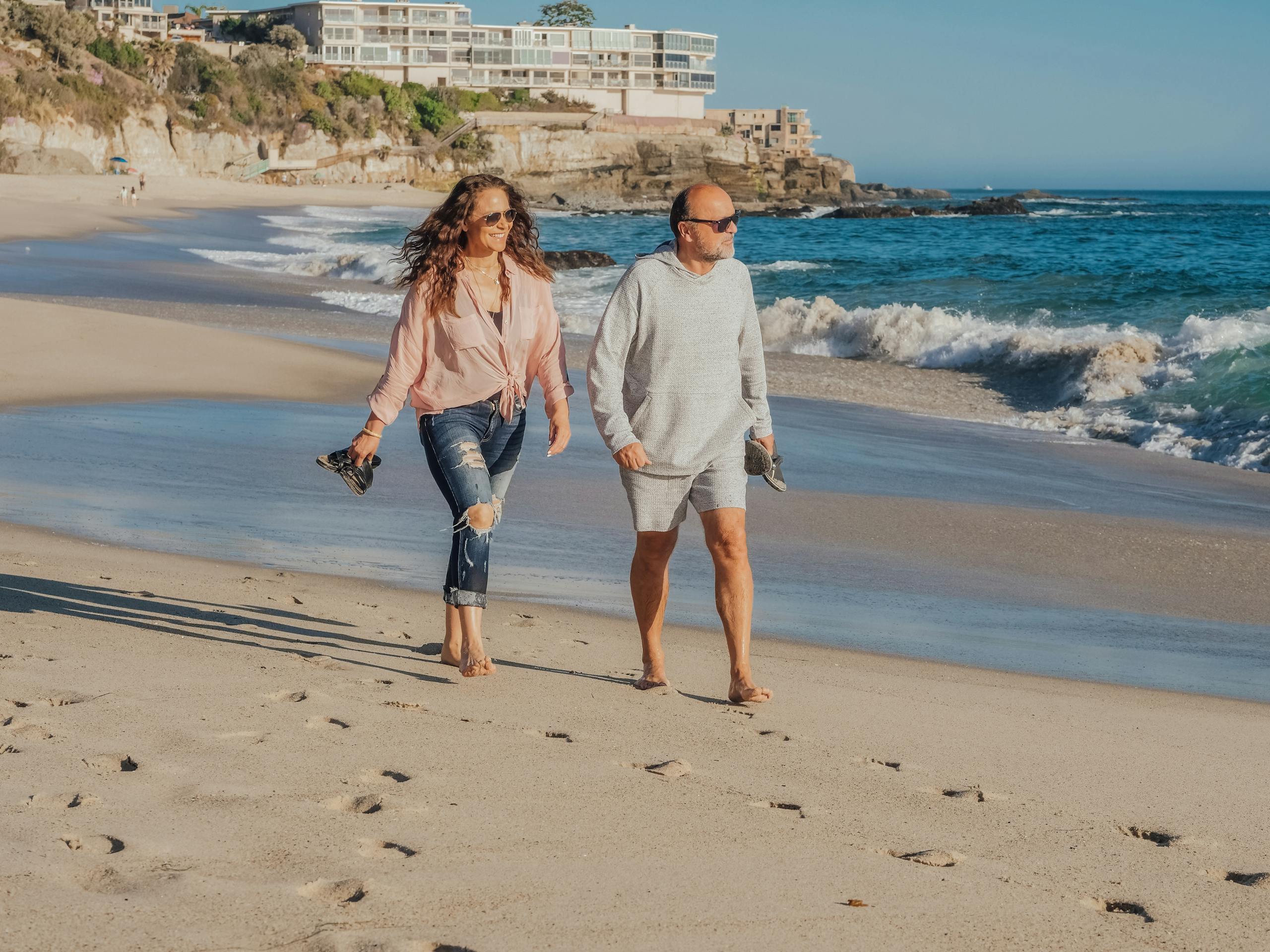 Elderly couple walking barefoot on a sunny beach by the coast, enjoying summer vacation.