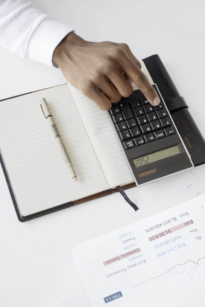 A hand using a calculator with financial documents and notebook, depicting an office setup for financial analysis.